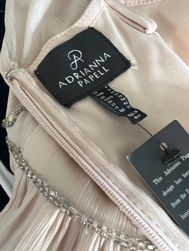 Puede incluir: Un vestido rosa p&aacute;lido con escote de cuentas y cremallera. El vestido tiene una etiqueta que dice "ADRIANNA PAPELL". El vestido est&aacute; hecho de una tela suave y fluida, talla 40.