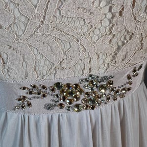 May include: A light pink dress with a lace bodice and a flowing skirt. The waist is accented with a decorative band featuring an arrangement of sparkling gemstones in shades of gold and silver.