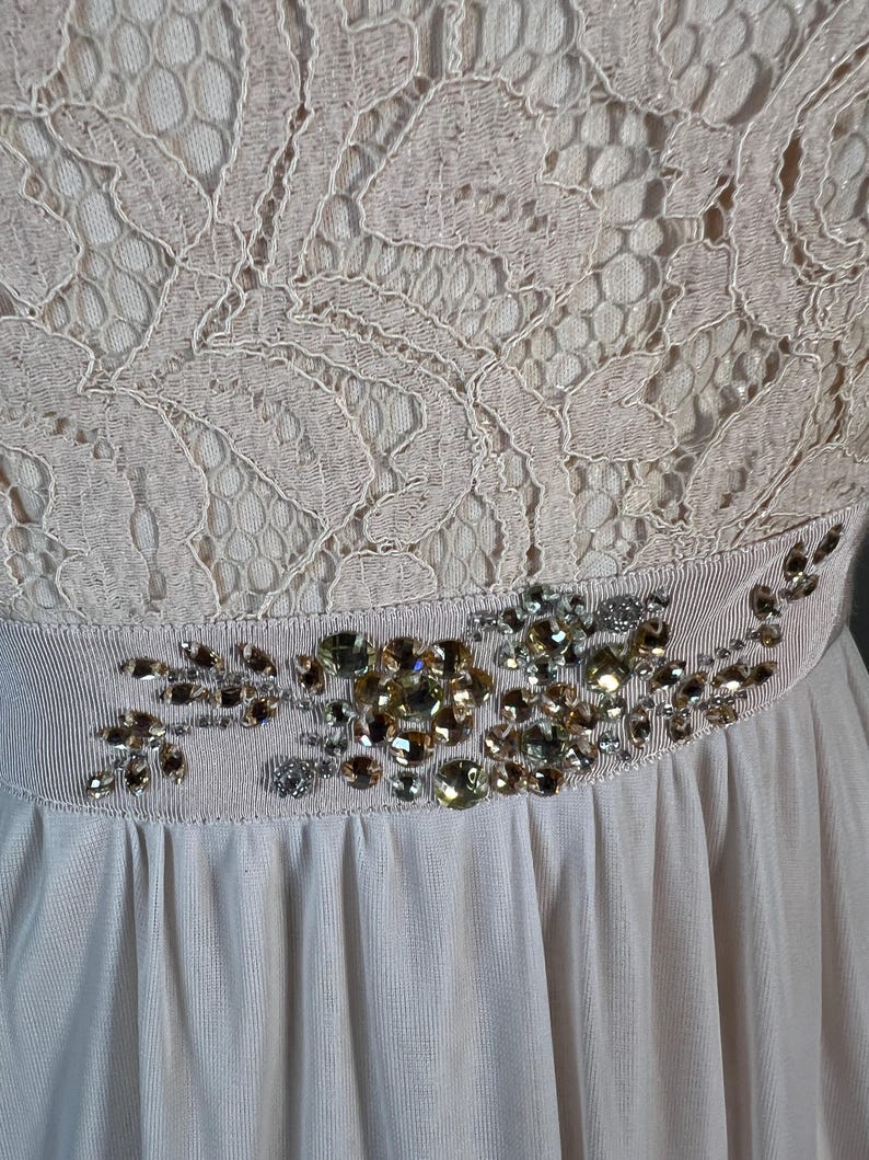 May include: A light pink dress with a lace bodice and a flowing skirt. The waist is accented with a decorative band featuring an arrangement of sparkling gemstones in shades of gold and silver.