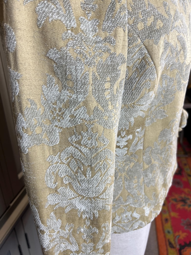 May include: Close-up of a beige jacket with a floral pattern in silver. The jacket has a tailored fit with a structured design. The fabric has a textured appearance, and the sleeves are visible.