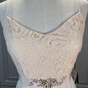 May include: A light pink formal dress with a V-neckline and spaghetti straps. The bodice is made of lace, and the waist is accented with a beaded embellishment. The skirt is a flowing, light pink fabric.
