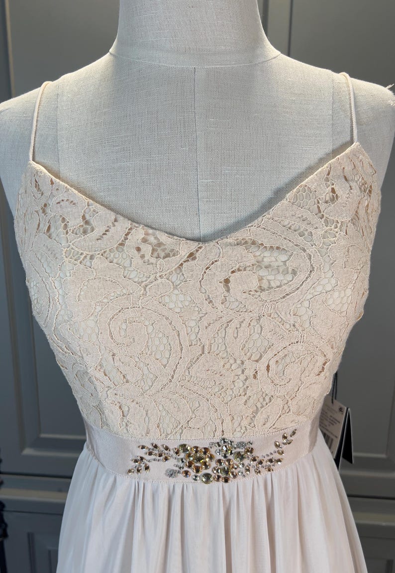 May include: A light pink formal dress with a V-neckline and spaghetti straps. The bodice is made of lace, and the waist is accented with a beaded embellishment. The skirt is a flowing, light pink fabric.