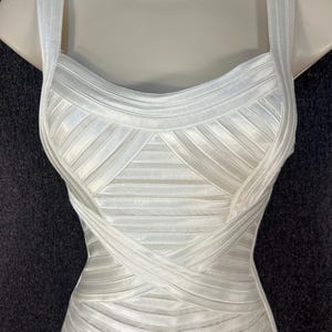 May include: A white, form-fitting dress with a structured bodice and thick straps. The dress features a ribbed design with crisscross detailing across the midsection. The dress is displayed on a mannequin.