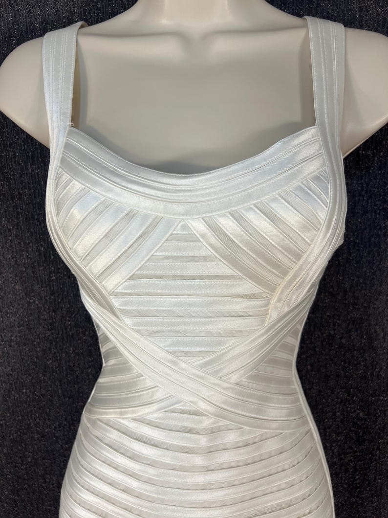 May include: A white, form-fitting dress with a structured bodice and thick straps. The dress features a ribbed design with crisscross detailing across the midsection. The dress is displayed on a mannequin.