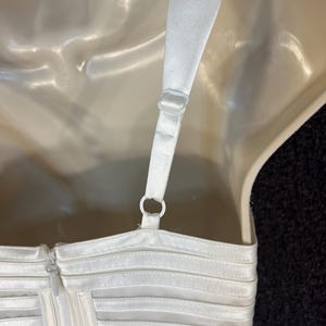 May include: Close-up of a white dress bodice with a zipper and adjustable straps. The dress features horizontal, sheer, pleated detailing. The straps are attached with plastic rings. The dress is displayed on a mannequin.