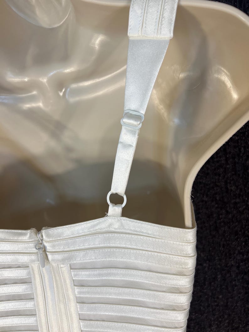 May include: Close-up of a white dress bodice with a zipper and adjustable straps. The dress features horizontal, sheer, pleated detailing. The straps are attached with plastic rings. The dress is displayed on a mannequin.