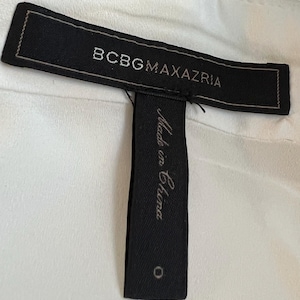 May include: A close-up of a black clothing tag with the brand name "BCBGMAXAZRIA" in white lettering. Below the brand name, a smaller tag reads "Made in China" in cursive. The background is a white fabric.