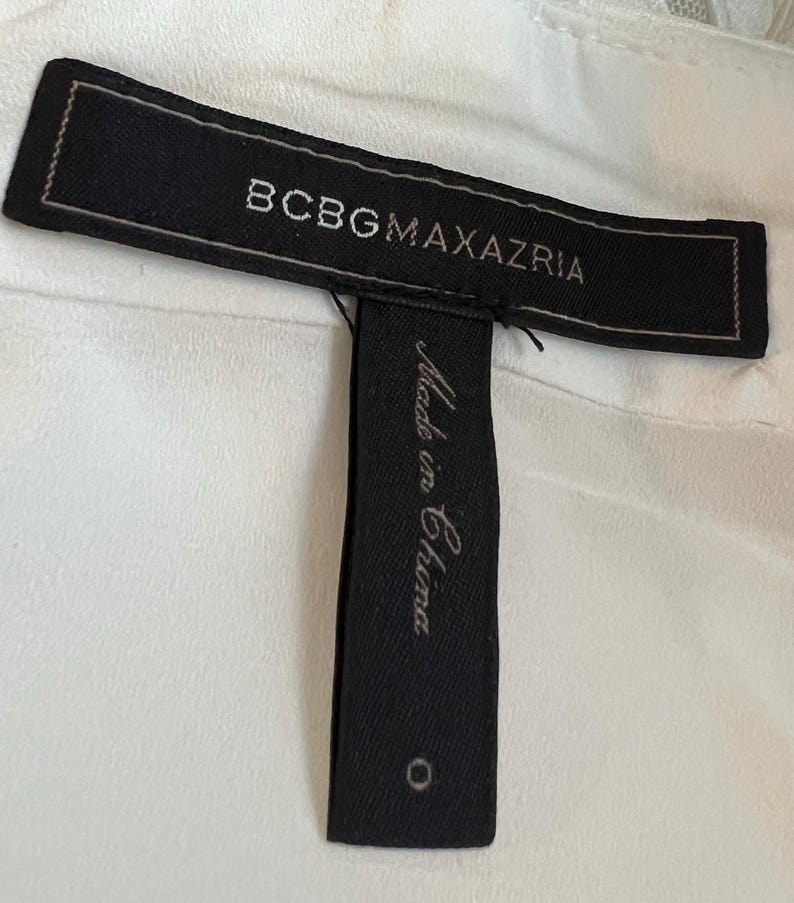 May include: A close-up of a black clothing tag with the brand name "BCBGMAXAZRIA" in white lettering. Below the brand name, a smaller tag reads "Made in China" in cursive. The background is a white fabric.