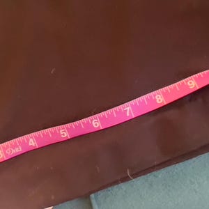 Puede incluir: Una tela marr&oacute;n oscuro con una cinta m&eacute;trica rosa colocada encima. La cinta muestra medidas de 2,5 a 30,5 cm. La tela parece ser una prenda de vestir o un art&iacute;culo similar.