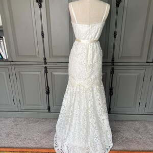 May include: A white lace wedding dress with a fitted bodice and a flowing skirt. The dress has a sweetheart neckline and a delicate lace pattern. The dress is accented with a satin ribbon at the waist.