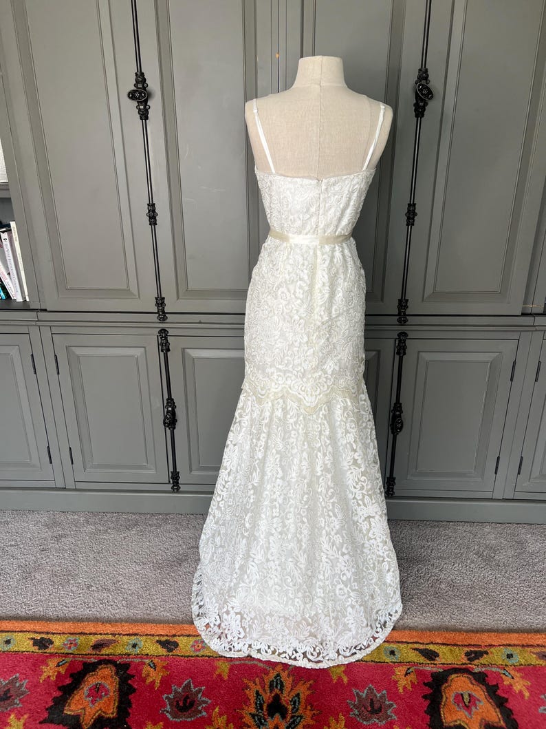 May include: A white lace wedding dress with a fitted bodice and a flowing skirt. The dress has a sweetheart neckline and a delicate lace pattern. The dress is accented with a satin ribbon at the waist.