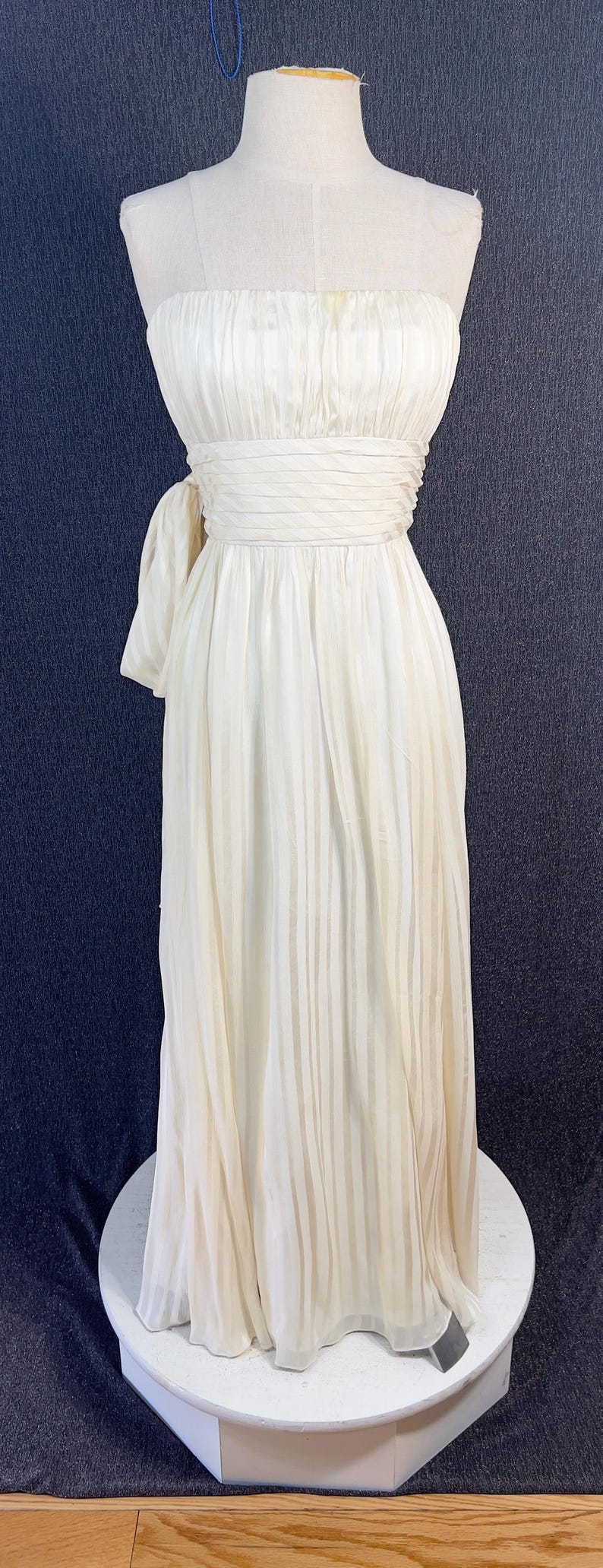May include: A floor-length, off-white strapless gown featuring a pleated bodice and a wide, horizontally pleated waistband. The skirt is made of a sheer, vertically striped fabric that flows to the floor, with a decorative fabric tie at the hip.