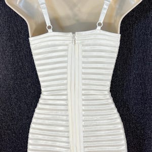 May include: A white, horizontally striped dress with thin straps and a zipper down the back. The dress is displayed on a mannequin and hangs from a hanger. The background is a dark, textured surface.