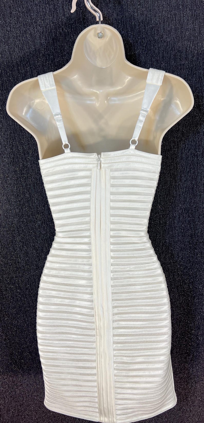 May include: A white, horizontally striped dress with thin straps and a zipper down the back. The dress is displayed on a mannequin and hangs from a hanger. The background is a dark, textured surface.