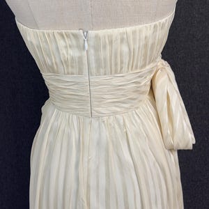 May include: A strapless cream-colored dress featuring a subtle vertical striped pattern. The bodice includes a gathered, ruched waistline and a side-tied bow detail. A vertical zipper closure is visible at the center back of the garment.