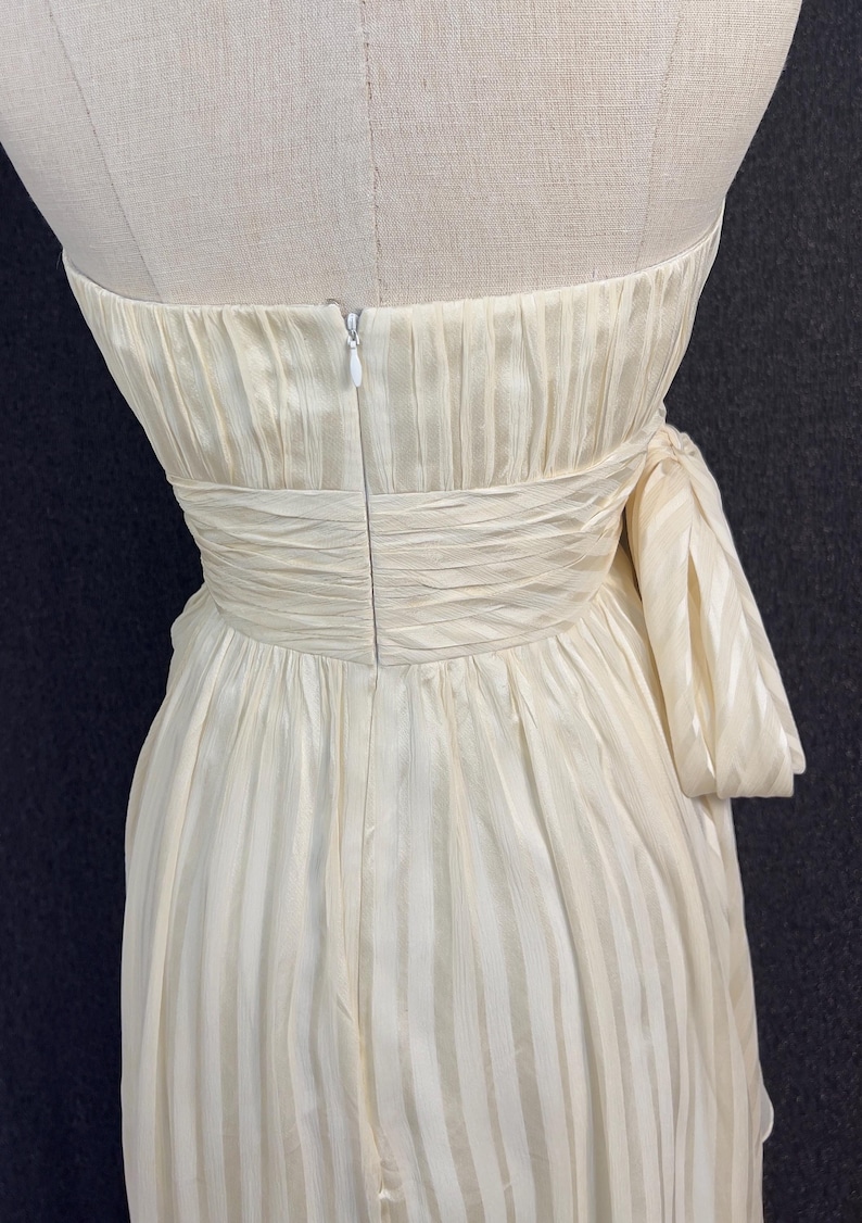 May include: A strapless cream-colored dress featuring a subtle vertical striped pattern. The bodice includes a gathered, ruched waistline and a side-tied bow detail. A vertical zipper closure is visible at the center back of the garment.