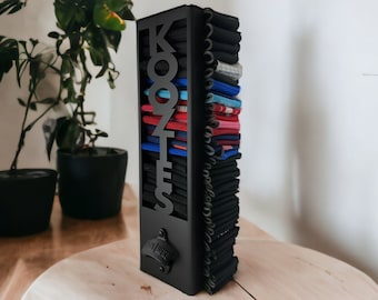 May include: Black metal wall-mounted organizer with the word "KOZTS" in large letters. The organizer has multiple slots for storing items, such as socks, ties, or belts. The organizer has a bottle opener attached to the bottom.