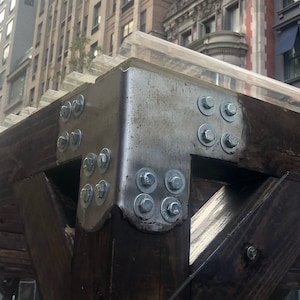 May include: A close-up of a wooden structure with metal brackets and bolts. The structure is covered with a clear plastic roof. The structure is in front of a tall building with many windows.