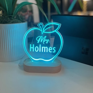 May include: A blue illuminated acrylic apple night light with the text "Mrs. Holmes" on it. The light is on a wooden base.