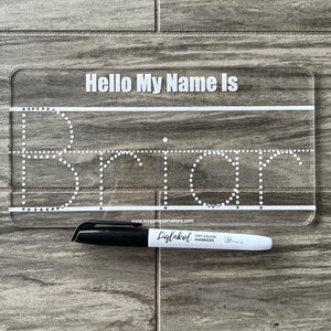 Pode incluir: Um quadro branco de pl&aacute;stico transparente com o texto "Hello My Name Is" e um contorno pontilhado da palavra "Brian". Um marcador de quadro branco preto est&aacute; deitado ao lado do quadro.