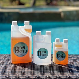Pode incluir: Três garrafas de plástico transparente com um design de bico duplo. As garrafas estão cheias de um líquido âmbar claro. As garrafas são etiquetadas com "Booze Bottle" com o endereço do site e o tamanho da garrafa em onças. As garrafas estão sobre uma superfície de vime marrom.