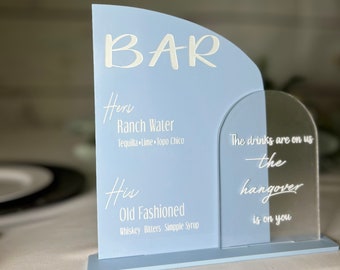 May include: A blue acrylic sign with the word "BAR" at the top. The sign lists two drink options: "Hers Ranch Water Tequila Lime Topo Chico" and "His Old Fashioned Whiskey Bitters Simple Syrup". The sign also includes the text "The drinks are on us - the hangover is on you".