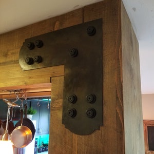 May include: A black metal corner brace with decorative detailing and multiple bolts, attached to a wooden beam.