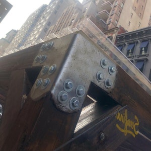 Pode incluir: Close-up de um suporte de metal com v&aacute;rios parafusos e porcas, fixado a uma viga de madeira. O suporte &eacute; pintado de prata e a viga &eacute; marrom. O fundo mostra um horizonte urbano com edif&iacute;cios.
