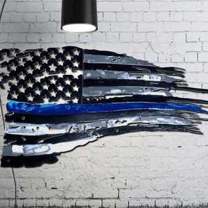May include: A distressed American flag with a blue line running horizontally across the center. The flag is made of metal and has a weathered, rustic look.