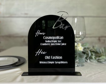 May include: Black acrylic sign with a rounded arch top. The sign says "Bar" at the top and lists two drink options: "Hers Cosmopolitan Vodka Triple Sec Cranberry juice/Lime juice" and "His Old Fashion Whiskey/Simple Syrup/Bitters".