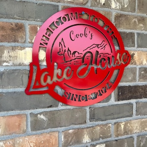 May include: Red metal sign with the text "Welcome to the Cook's Lake House Since 2021". The sign features a stylized lake scene with trees and a house.