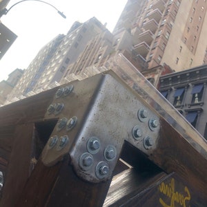 May include: Close-up of a metal bracket with multiple bolts and nuts securing a wooden beam. The bracket is attached to a clear plastic panel. The background shows a city skyline with buildings.