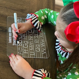 Op de afbeelding: Een doorzichtig acryl traceerbord met de tekst "Learn my ABCs Trace" en het alfabet. Een kind gebruikt een zwarte stift om de letters te traceren. Het kind draagt een groen polkadot shirt met zebra gestreepte manchetten.