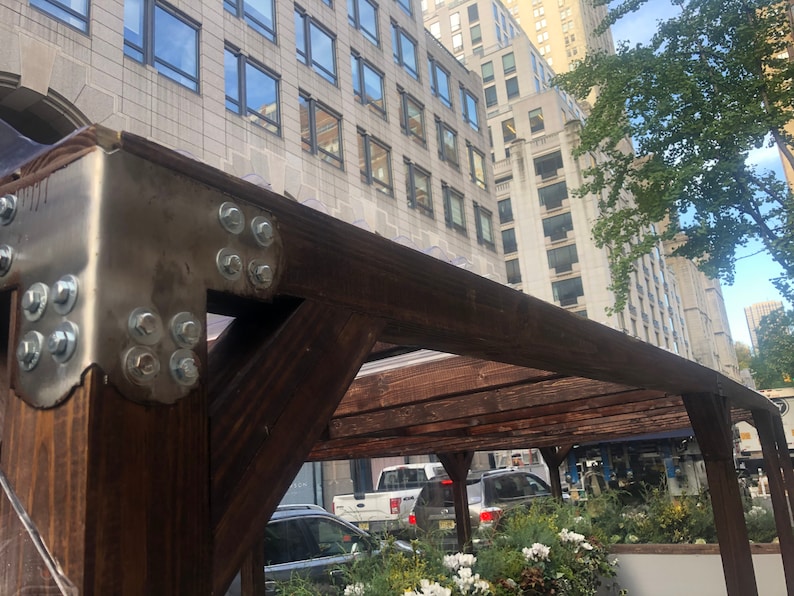 Pode incluir: Uma p&eacute;rgola de madeira com um suporte de metal fixado com v&aacute;rios parafusos. A p&eacute;rgola &eacute; feita de madeira marrom escura e est&aacute; presa a um pr&eacute;dio com grandes janelas.