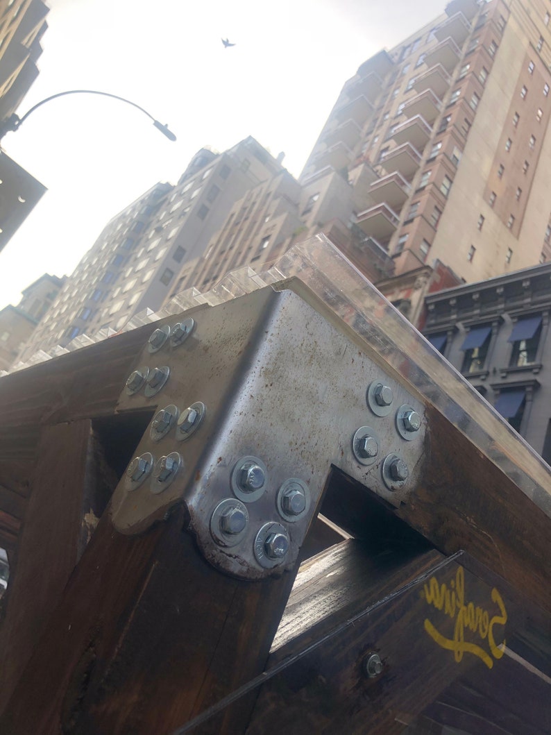 Pode incluir: Close-up de um suporte de metal com v&aacute;rios parafusos e porcas, fixado a uma viga de madeira. O suporte &eacute; pintado de prata e a viga &eacute; marrom. O fundo mostra um horizonte urbano com edif&iacute;cios.