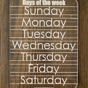 Op de afbeelding: Transparant acryl leerbord voor de dagen van de week met witte letters. De woorden "Days of the week" staan bovenaan, gevolgd door de dagen van de week. Stippellijnen zijn inbegrepen voor schrijfoefeningen. De website www.texasmetalmakers.com staat onderaan.