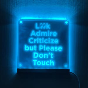 May include: A blue neon sign with the text "Look Admire Criticize but Please Don't Touch" on a white background.