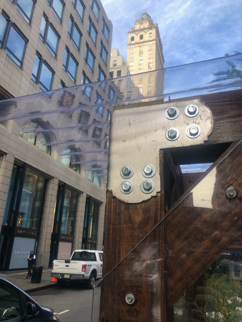 May include: A close-up of a wooden structure with a clear plastic roof. The structure is attached to a building with metal brackets and screws. The building is made of brick and has large windows.