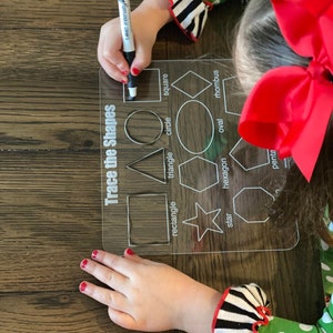 Op de afbeelding: Een doorzichtig acryl traceerbord met de tekst "Trace the Shapes" en diverse geometrische vormen. Een kinderhand, die een zwarte stift vasthoudt, trekt een vierkant over. Het kind draagt een groene jurk met witte stippen en zwart-wit gestreepte mouwen.
