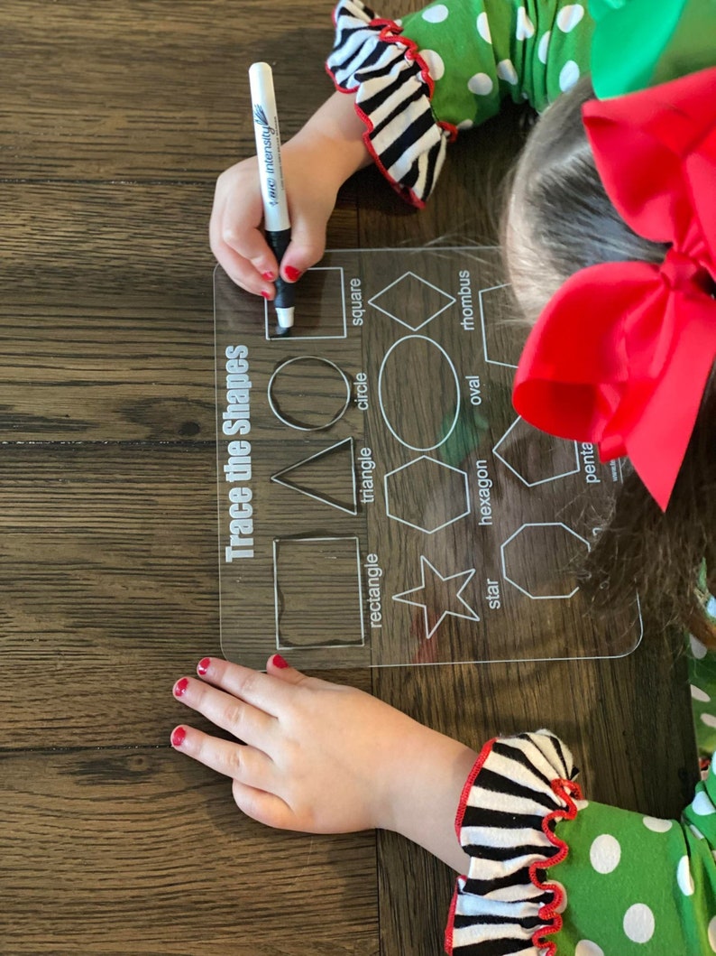 Op de afbeelding: Een doorzichtig acryl traceerbord met de tekst "Trace the Shapes" en diverse geometrische vormen. Een kinderhand, die een zwarte stift vasthoudt, trekt een vierkant over. Het kind draagt een groene jurk met witte stippen en zwart-wit gestreepte mouwen.