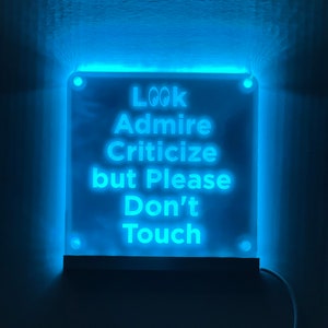 May include: A white sign with blue neon lighting, mounted on a wall. The sign reads "Look Admire Criticize but Please Don't Touch".