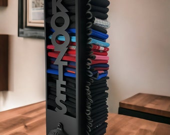 May include: Black metal can koozie holder with the word "KOZIES" cut out. The holder has a bottle opener attached to the bottom. The holder is filled with a stack of colorful can koozies.