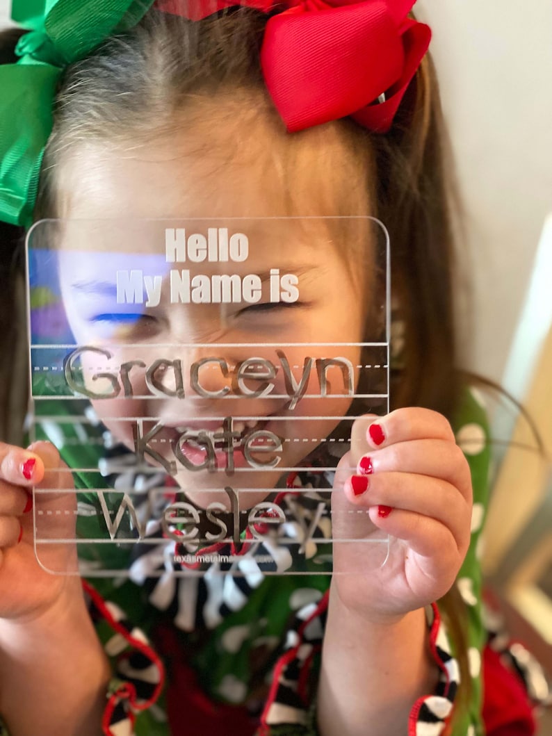 Op de afbeelding: Transparante acrylen naamplaat met de tekst "Hello My Name is" en drie regels voor namen. De namen Graceyn, Kate en Wesley zijn gegraveerd. De plaat wordt vastgehouden door een persoon met rode nagellak.