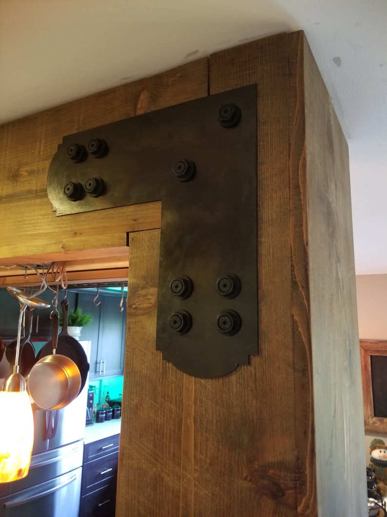 May include: A black metal corner brace with decorative detailing and multiple bolts, attached to a wooden beam.