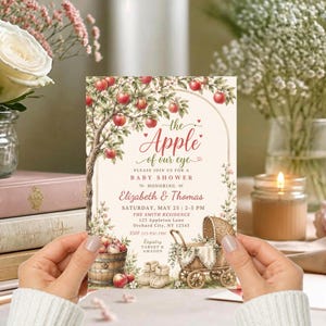 May include: A rectangular baby shower invitation card featuring a watercolour illustration of an apple tree, a wooden barrel filled with apples, and a vintage wicker baby carriage. The text reads: the Apple of our eye, Elizabeth & Thomas.