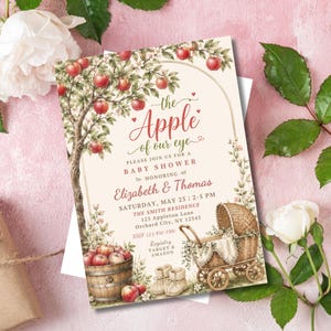 May include: A rectangular baby shower invitation card featuring a watercolour illustration of an apple tree, a wooden barrel filled with red apples, and a wicker baby carriage. The text reads: the Apple of our eye, PLEASE JOIN US FOR A BABY SHOWER, HONORING Elizabeth & Thomas, SATURDAY, MAY 25 | 2-5 PM, THE SMITH RESIDENCE, 123 Appleton Lane, Orchard City, NY 12345, RSVP 123-456-7890, Registry TARGET & AMAZON.