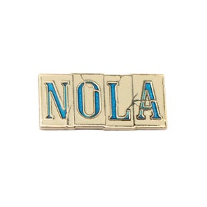 May include: A rectangular pin with the word "NOLA" in blue letters. The letters are set against a cream-colored background with a gold border. The pin has a distressed, vintage look.