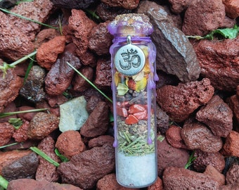 May include: A small glass bottle with a purple wax seal and a silver charm with the Om symbol. The bottle is filled with various herbs, spices, and crystals. The bottle is sitting on a bed of red lava rocks.