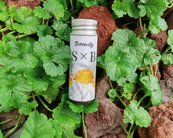 May include: A small silver bottle with a white label that says "Serenity S x B" and a yellow floral design. The bottle is sitting on a bed of green leaves and brown rocks.