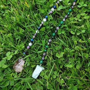May include: A crystal necklace with a variety of stones, including amethyst, blue tiger's eye, and malachite. The necklace has two charms, a pentagram and a circle with a dot in the center. The necklace is lying on a bed of green grass.